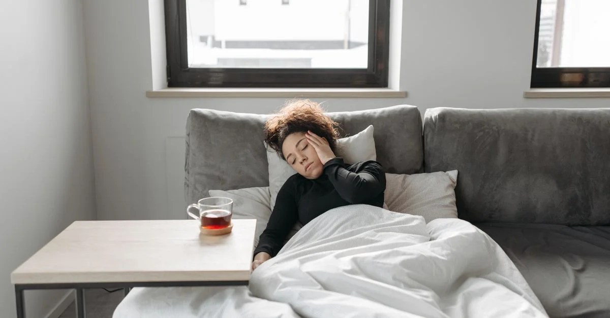 Une femme repose sur un canapé à l'intérieur, se sentant mal, avec une tasse de thé à proximité.