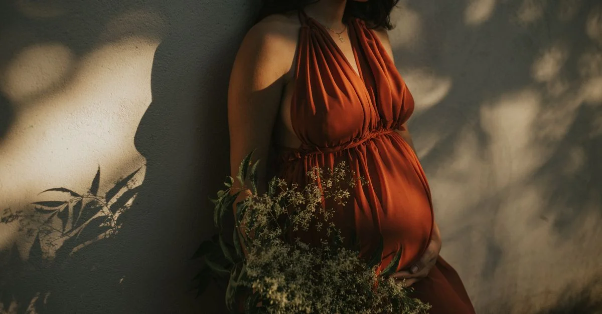 Une femme enceinte sereine dans une robe vibrante tient des fleurs contre un mur texturé.