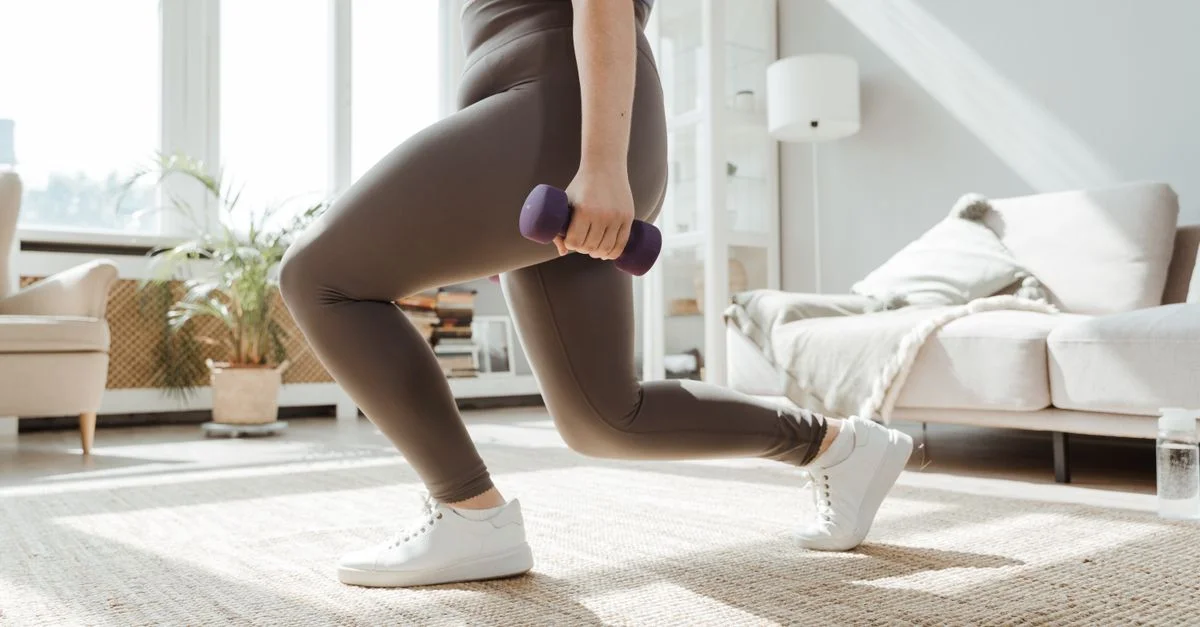 Une femme en tenue de sport effectue un exercice de fente avec des haltères à l'intérieur.