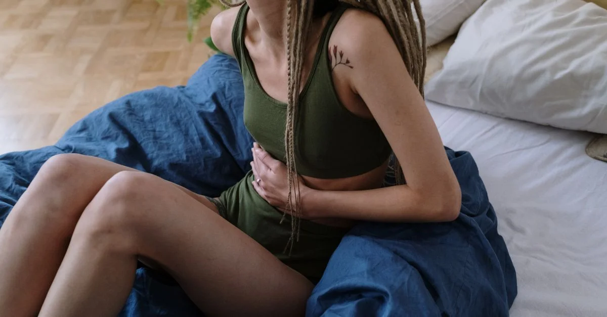 Femme assise sur le lit tenant son ventre, indiquant une douleur ou un inconfort, probablement dû à des crampes.