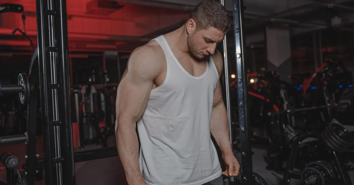 Un homme musclé dans une salle de sport portant un débardeur blanc, entouré d'équipements de musculation.