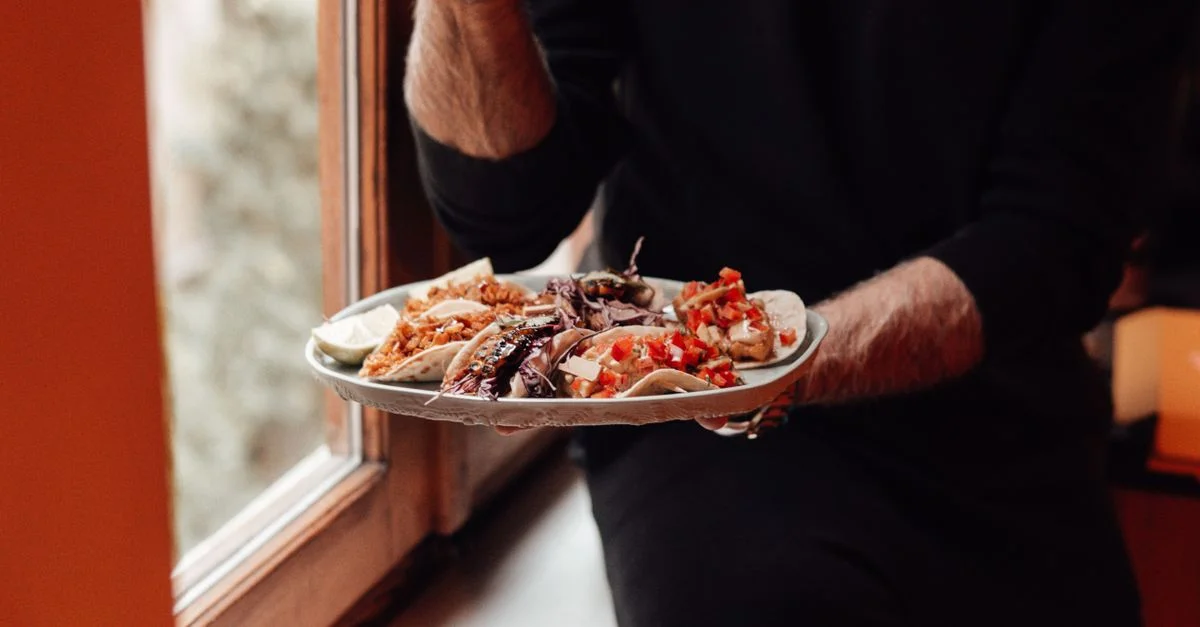 Une personne tenant une assiette de tacos variés près d'une fenêtre, profitant d'un repas frais à l'intérieur.