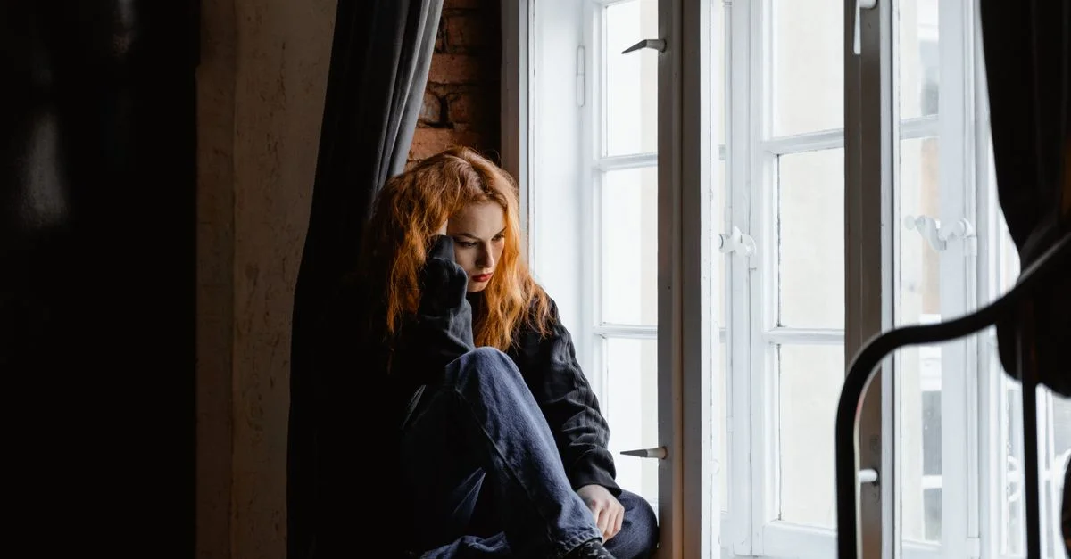 Une femme est assise près d'une fenêtre, perdue dans ses pensées profondes et avec une lumière moody.