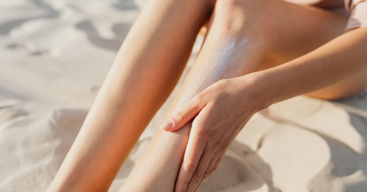 Gros plan d'une femme appliquant de la crème solaire sur ses jambes tout en étant assise sur le sable de la plage.
