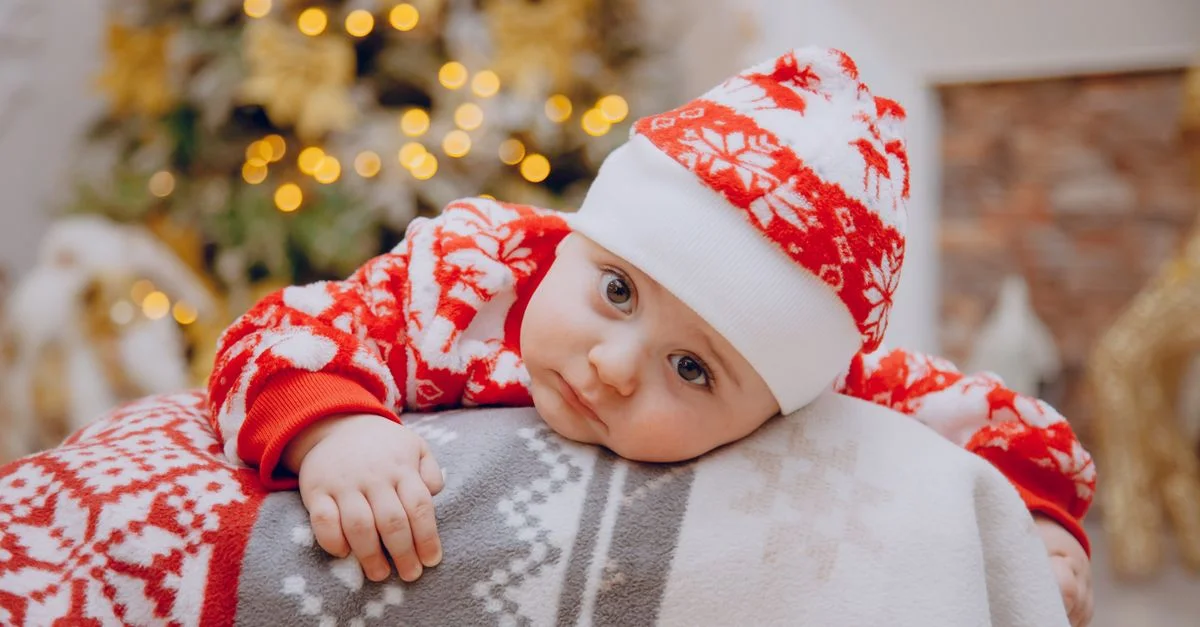 Charmant bébé en tenue rouge festive se reposant à côté d'un arbre de Noël décoré à l'intérieur.