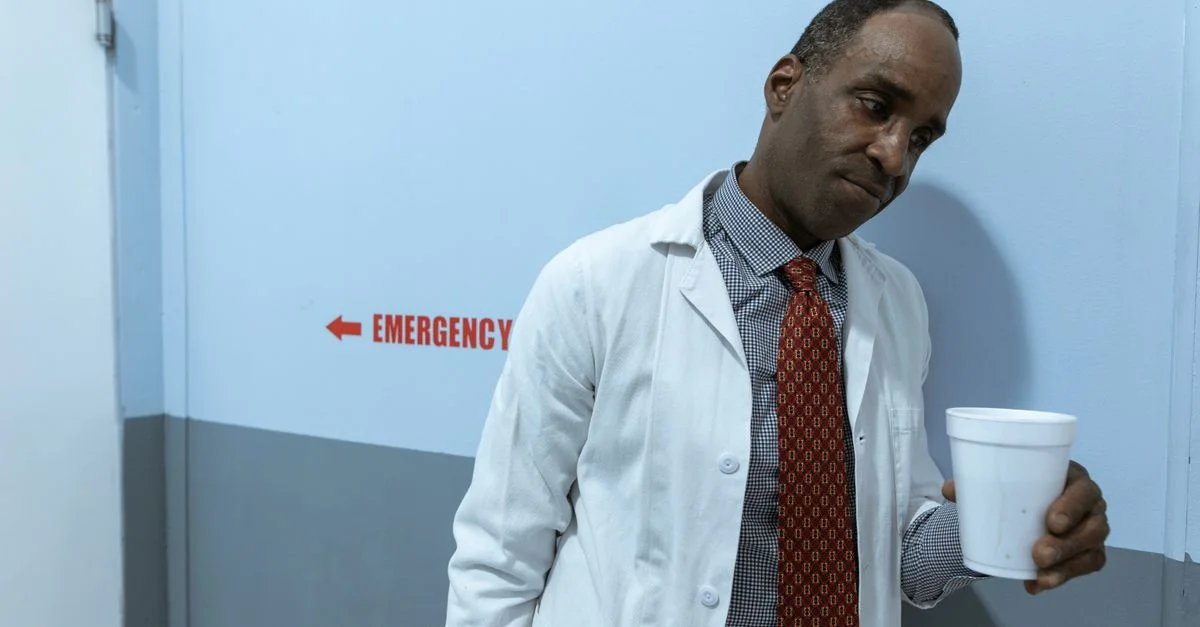 Un professionnel de la santé en blouse blanche tenant une tasse dans un couloir d'hôpital près de la section des urgences.