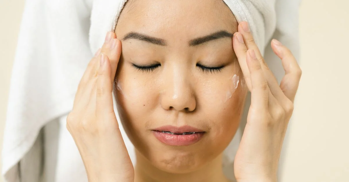 Femme asiatique avec une serviette sur la tête appliquant un hydratant pour sa routine de soin de la peau.