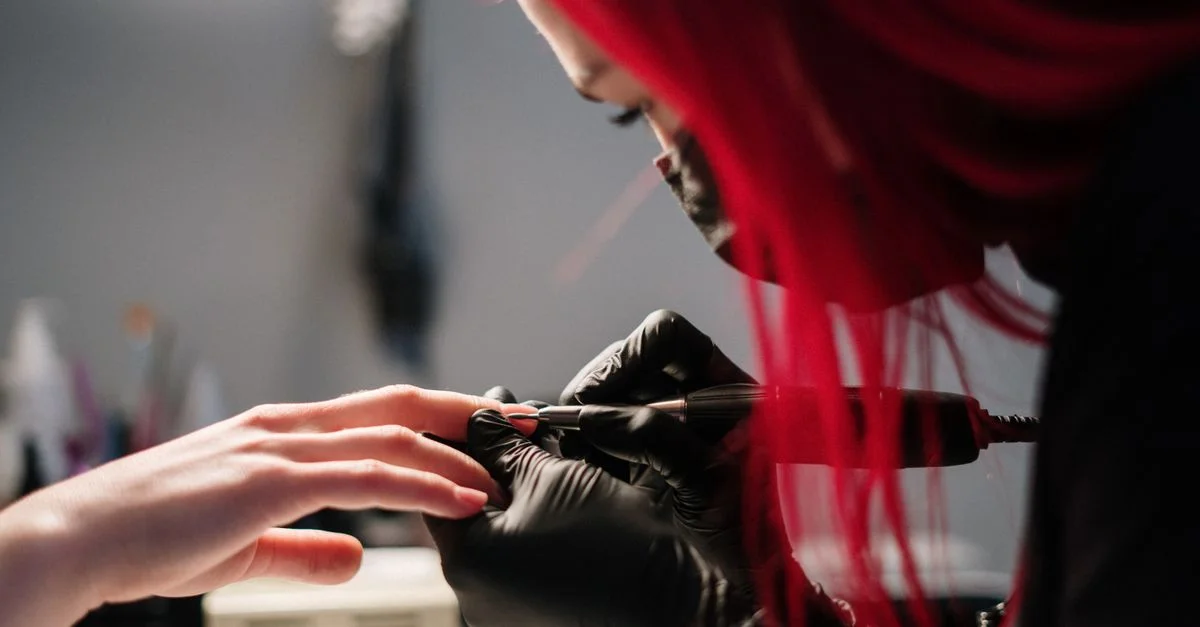 Une technicienne des ongles avec des cheveux rouges réalise une manucure en portant des gants noirs dans un salon.