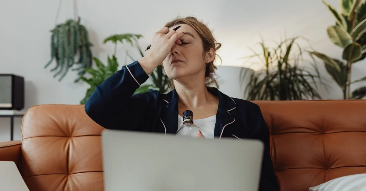 Femme stressée assise avec une boisson, un ordinateur portable et des plantes autour, exprimant de l'épuisement.
