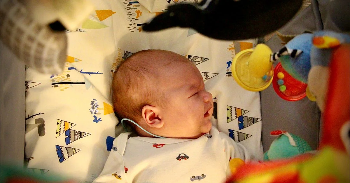 Bébé nouveau-né pleurant allongé dans un berceau entouré de jouets colorés dans un cadre douillet.