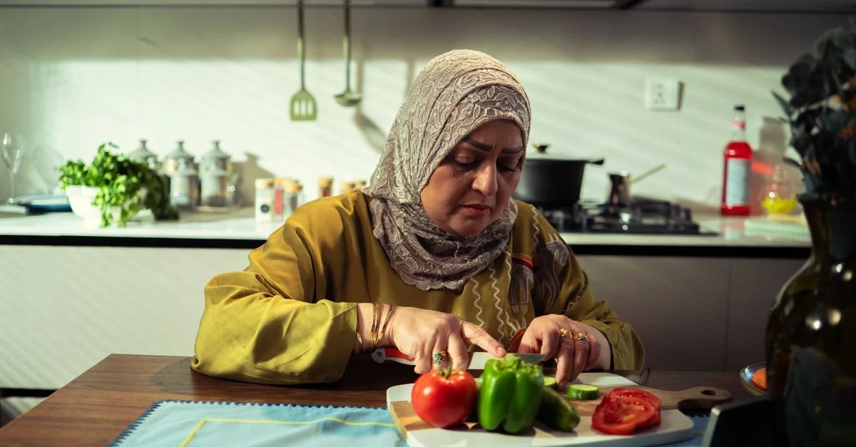 Une femme, concentrée sur la préparation de ses repas, coupe des poivrons verts et des tomates dans sa cuisine.
