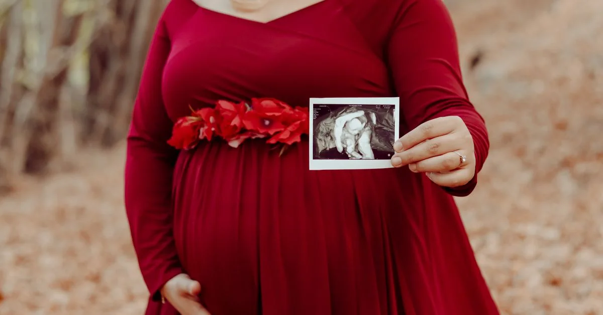 Femme enceinte en robe rouge montrant une échographie dans un cadre forestier automnal serein.