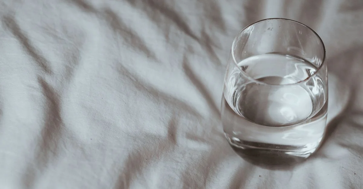 Prise de vue minimaliste d'un verre d'eau reposant sur une couette blanche, prise à l'intérieur.