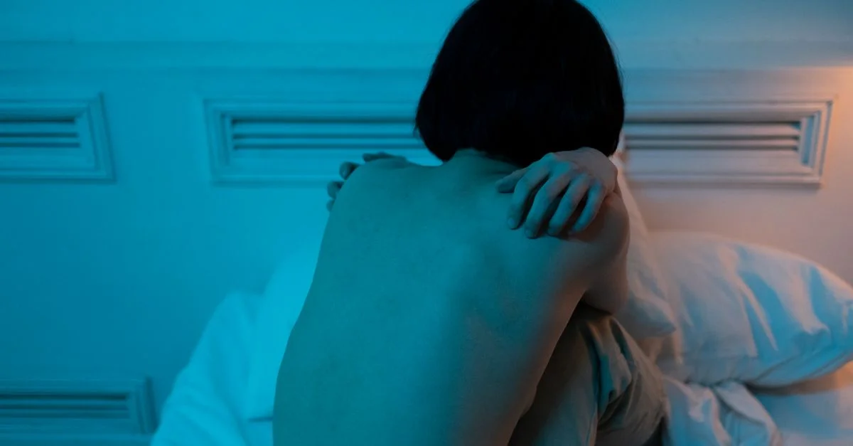 Une femme est assise seule sur un lit dans une pièce faiblement éclairée, exprimant la solitude.