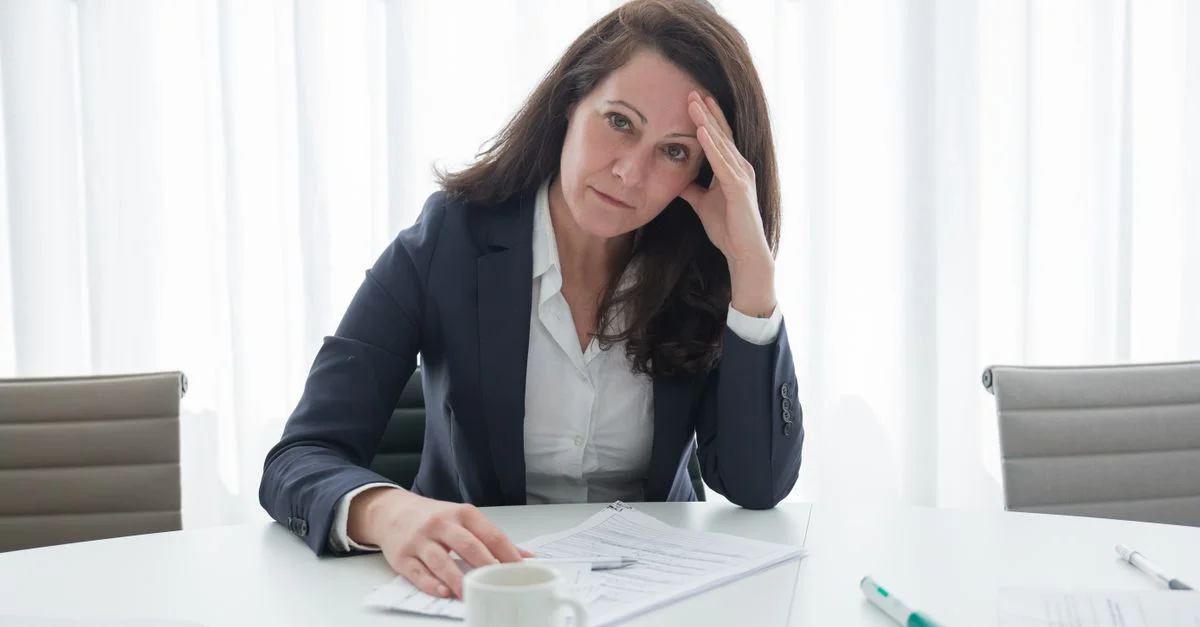 Une femme d'affaires stressée en tenue professionnelle est assise à un bureau avec des papiers, l'air accablé.