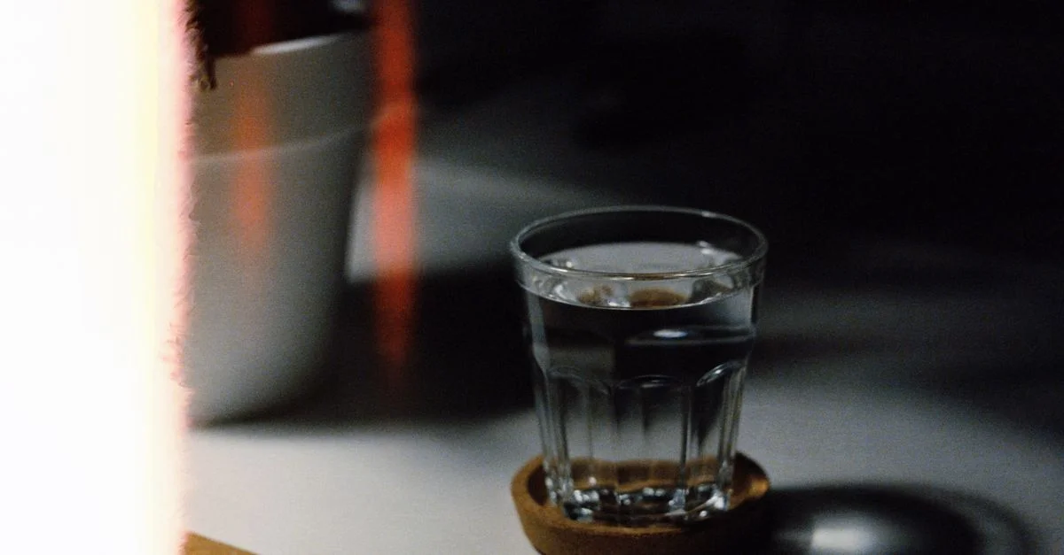 Une photo artistique et sombre d'un verre d'eau sur un dessous de verre à l'intérieur, mettant en valeur des ombres et un éclairage dramatiques.