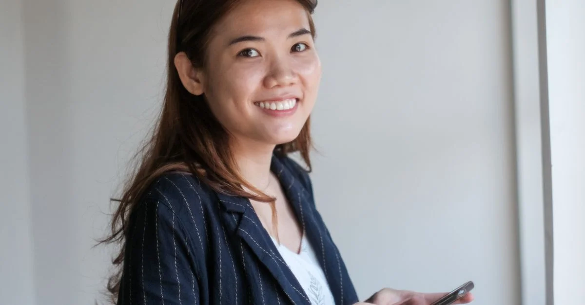 Femme souriante, debout à l'intérieur, tenant un smartphone, illustrant un mode de vie moderne et la connectivité.