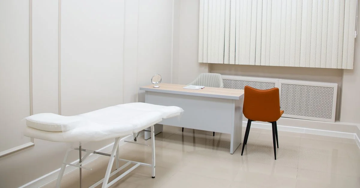 Une salle d'examen médical minimaliste avec un bureau, des chaises et un lit d'examen dans un cadre stérile et neutre.