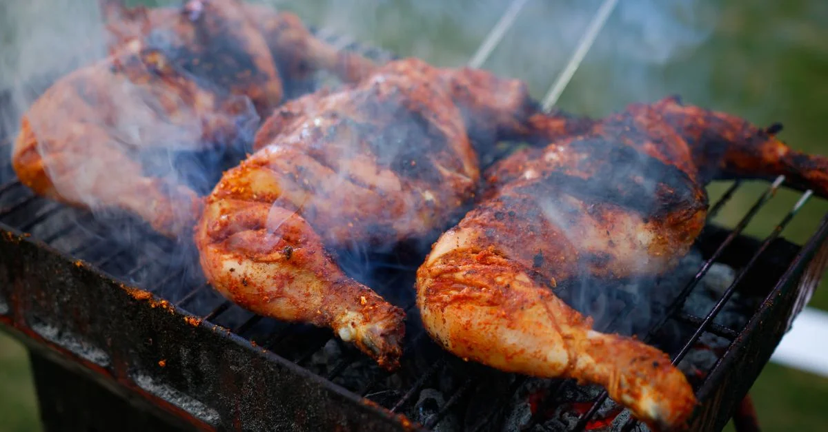 Gros plan sur du poulet épicé en train de griller sur un barbecue au charbon avec de la fumée visible en extérieur.