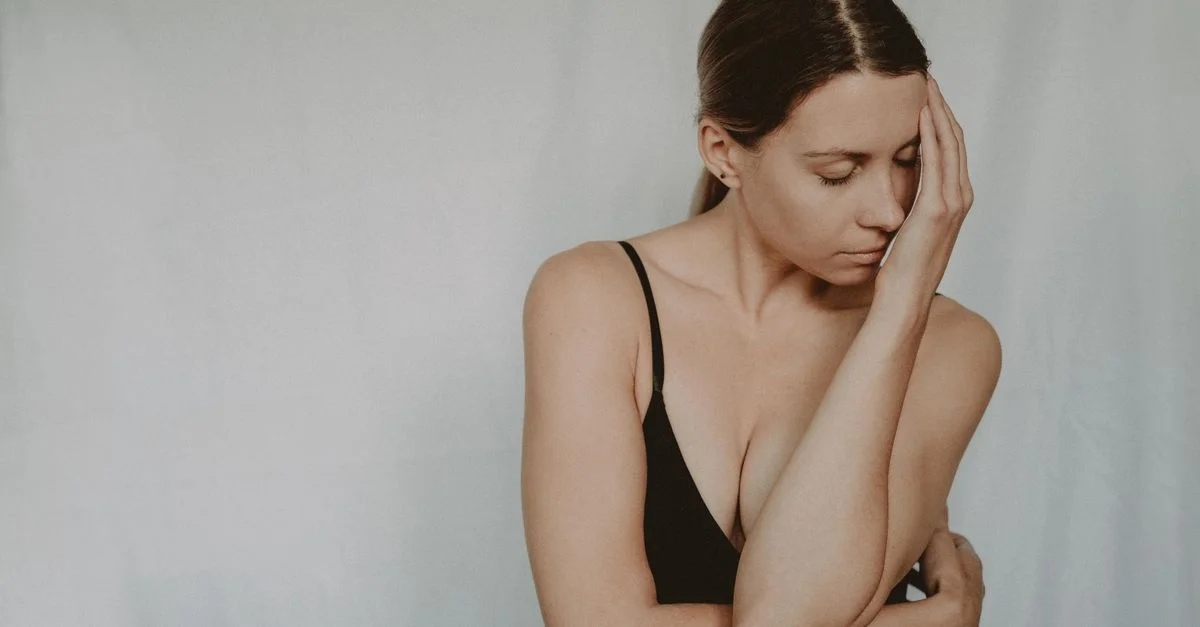 Femme en soutien-gorge noir avec une expression pensive, main sur le visage contre un fond gris.