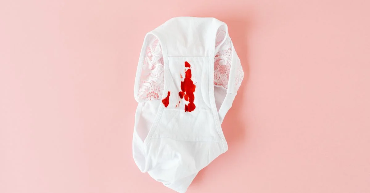 Culotte en dentelle blanche avec une tache menstruelle visible sur fond rose.
