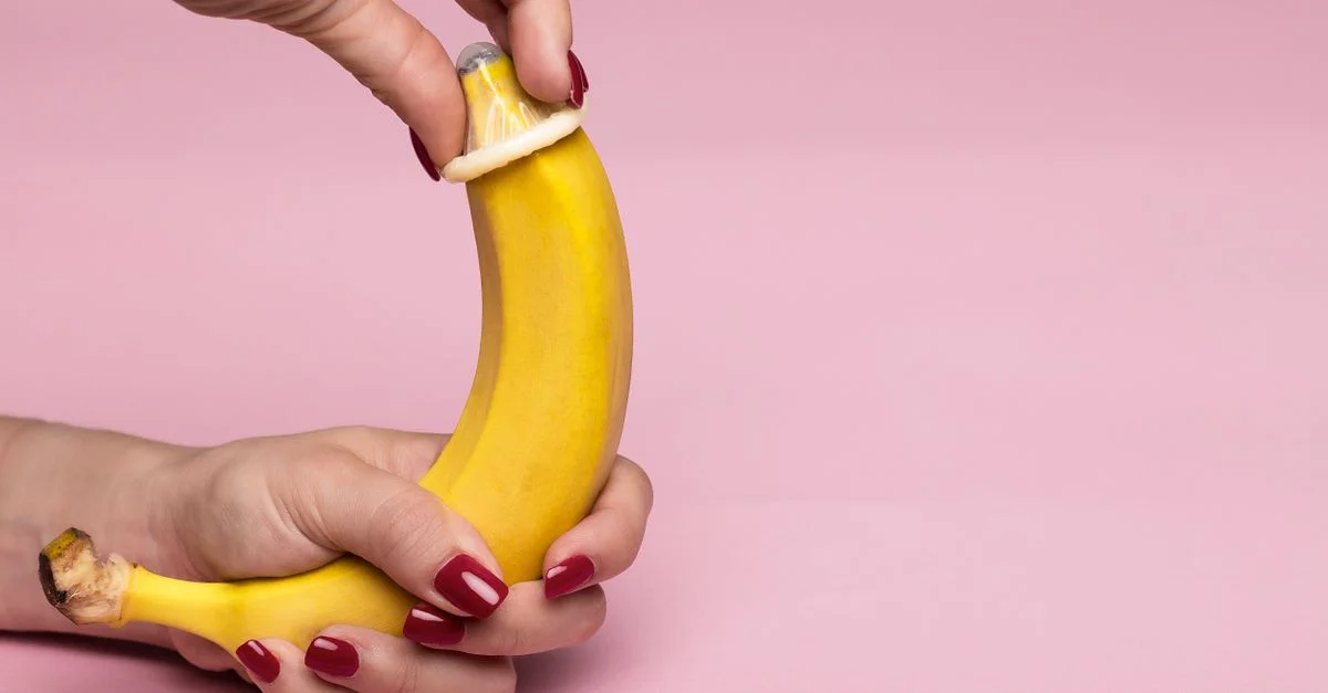 Gros plan de mains appliquant un préservatif sur une banane sur fond rose symbolisant le sexe sûr.