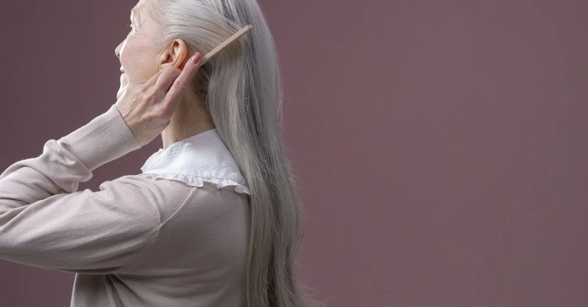 Femme âgée avec de longs cheveux gris se coiffant, sur un fond violet.