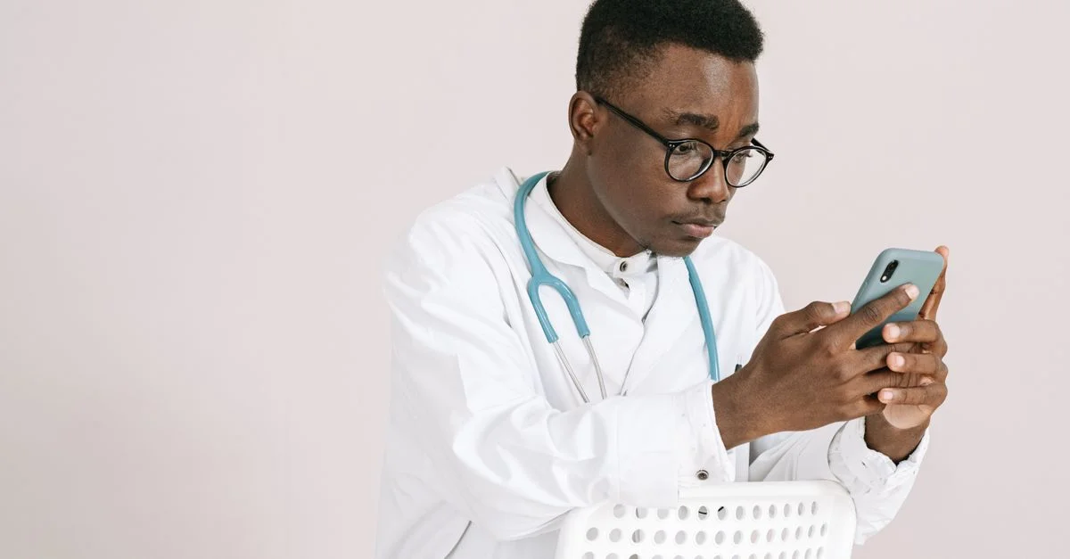 Jeune médecin masculin avec un stéthoscope parcourant son smartphone à l'intérieur.