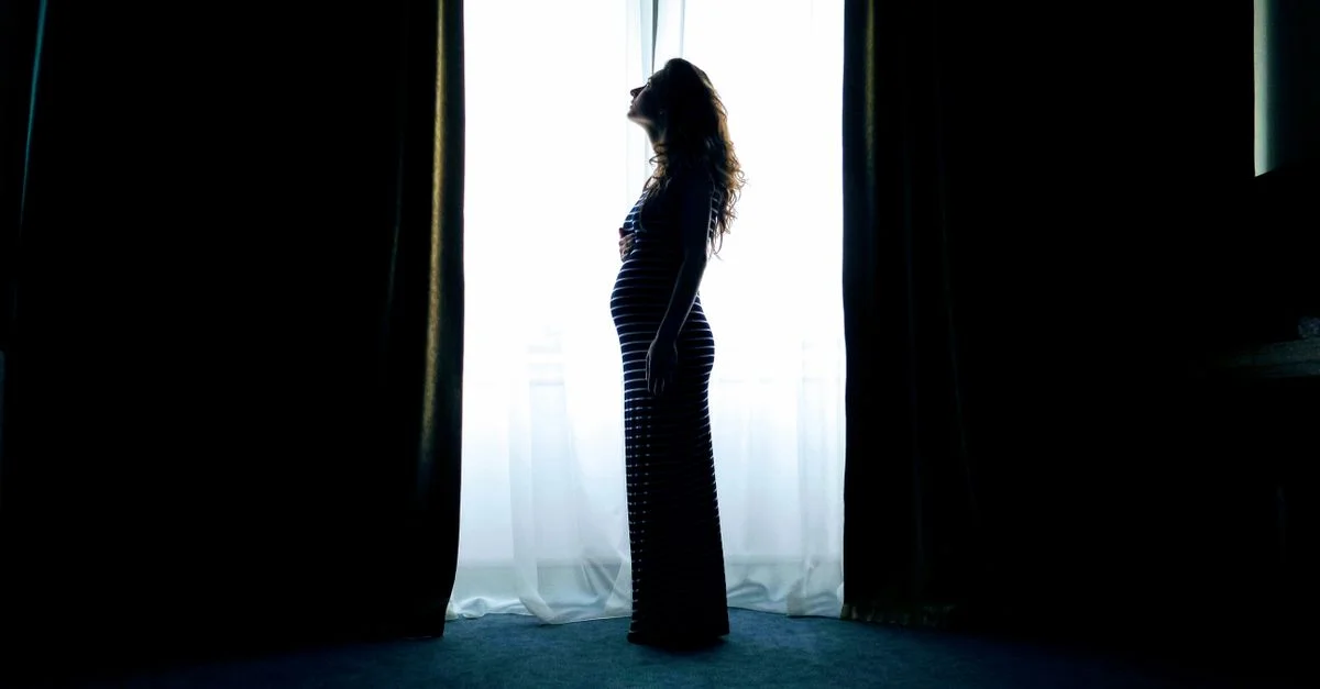 Silhouette d'une femme enceinte debout près d'une fenêtre lumineuse, capturée dans un cadre intérieur serein.
