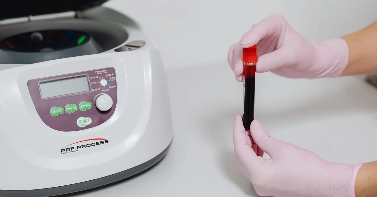 Gros plan de mains gantées tenant un échantillon de sang à côté d'une centrifugeuse.