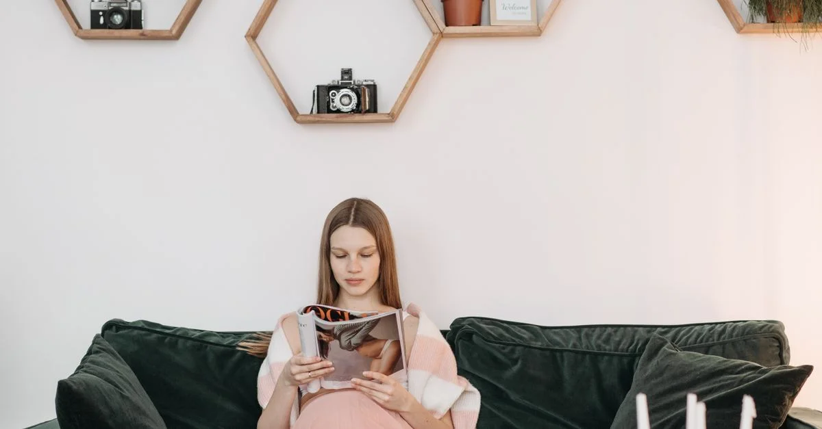 Une femme enceinte sereine lisant un magazine sur un canapé confortable, entourée d'étagères décoratives.