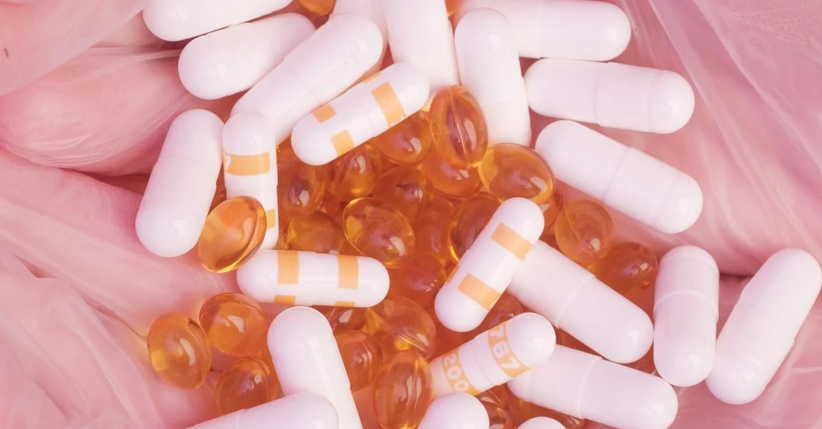 Gros plan sur un assortiment de gélules blanches et orange ainsi que des gélules en gel sur un doux fond rose texturé.