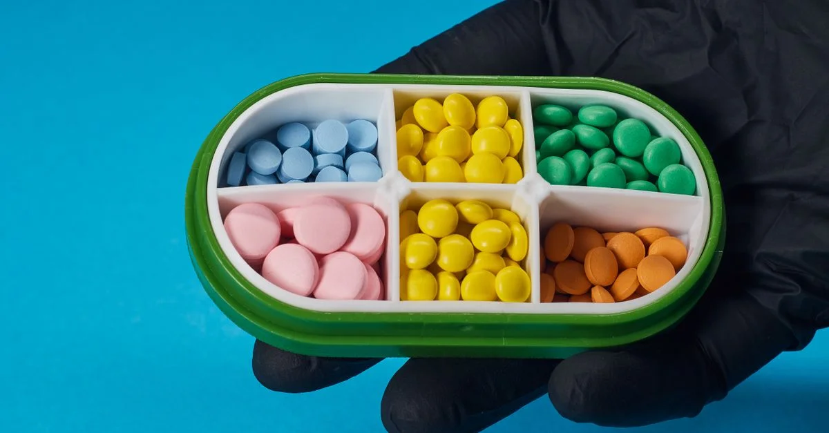 Des pilules colorées dans une boîte à quatre compartiments tenues par une main gantée sur un fond bleu.