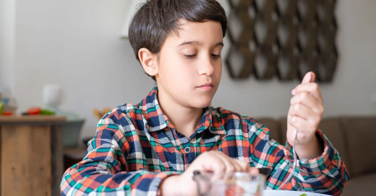 Un jeune garçon en chemise à carreaux déguste son petit déjeuner avec du jus et des ustensiles à une table familiale.