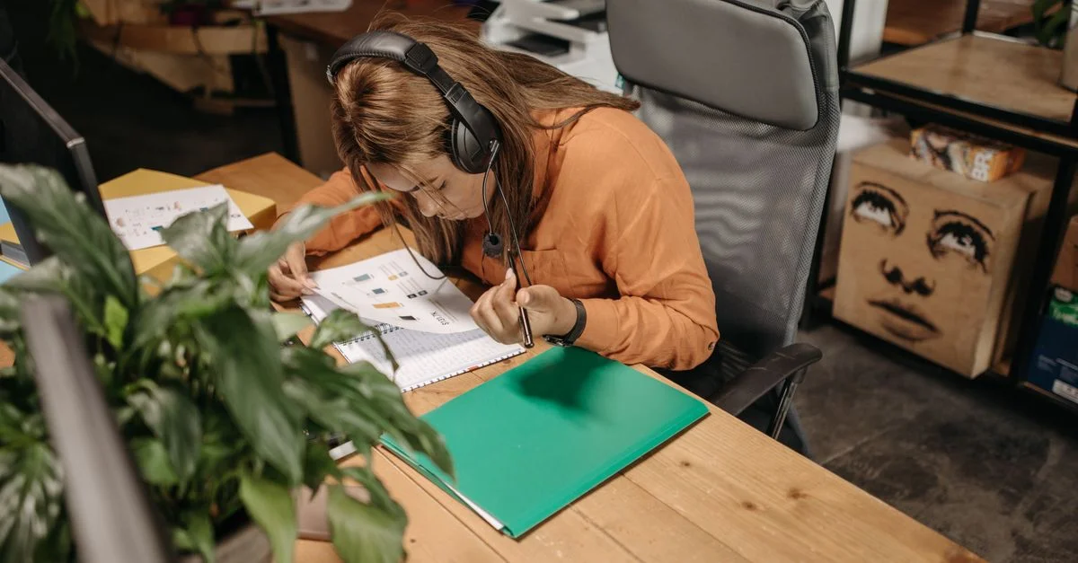 Une femme concentrée en chemise orange travaille assidûment à son bureau avec un casque, entourée d'objets de bureau.