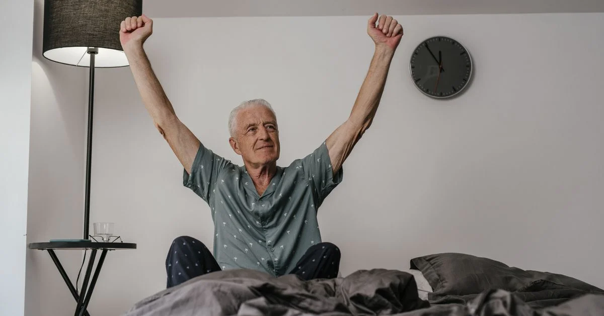 Homme âgé en pyjama se réveillant avec les bras étendus dans un cadre de chambre confortable.