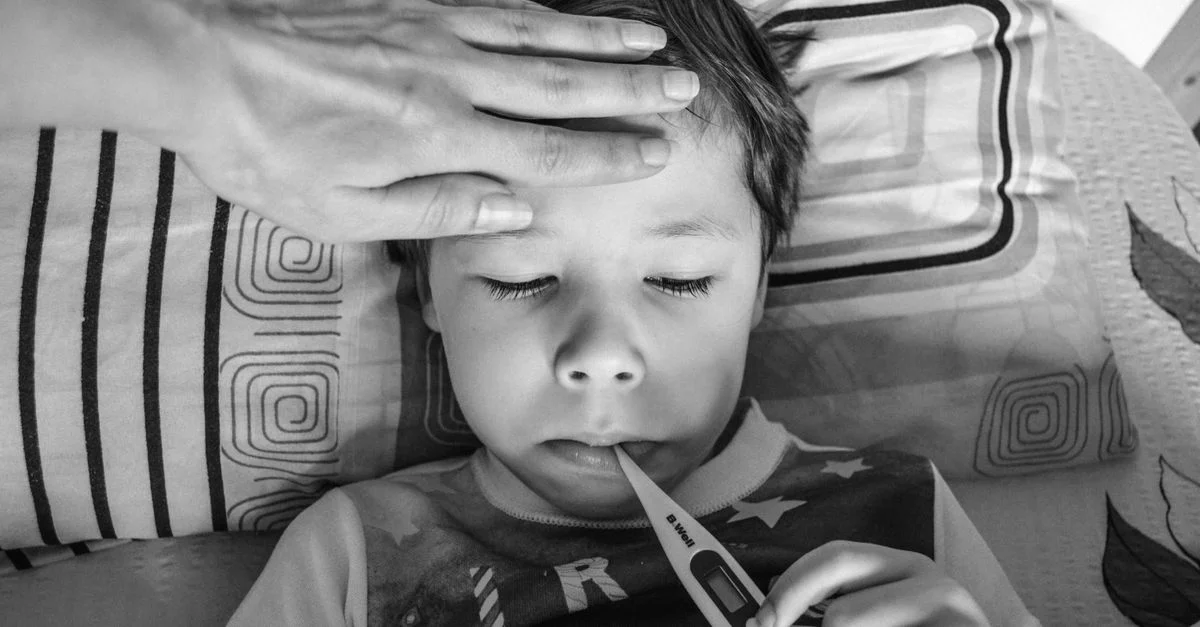 Un enfant se reposant avec un thermomètre, surveillé par une main bienveillante, à l'intérieur.