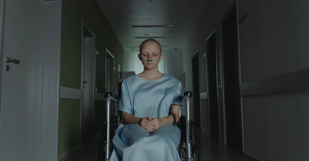 Femme en blouse d'hôpital assise dans un fauteuil roulant, perdue dans ses pensées, dans un couloir faiblement éclairé.