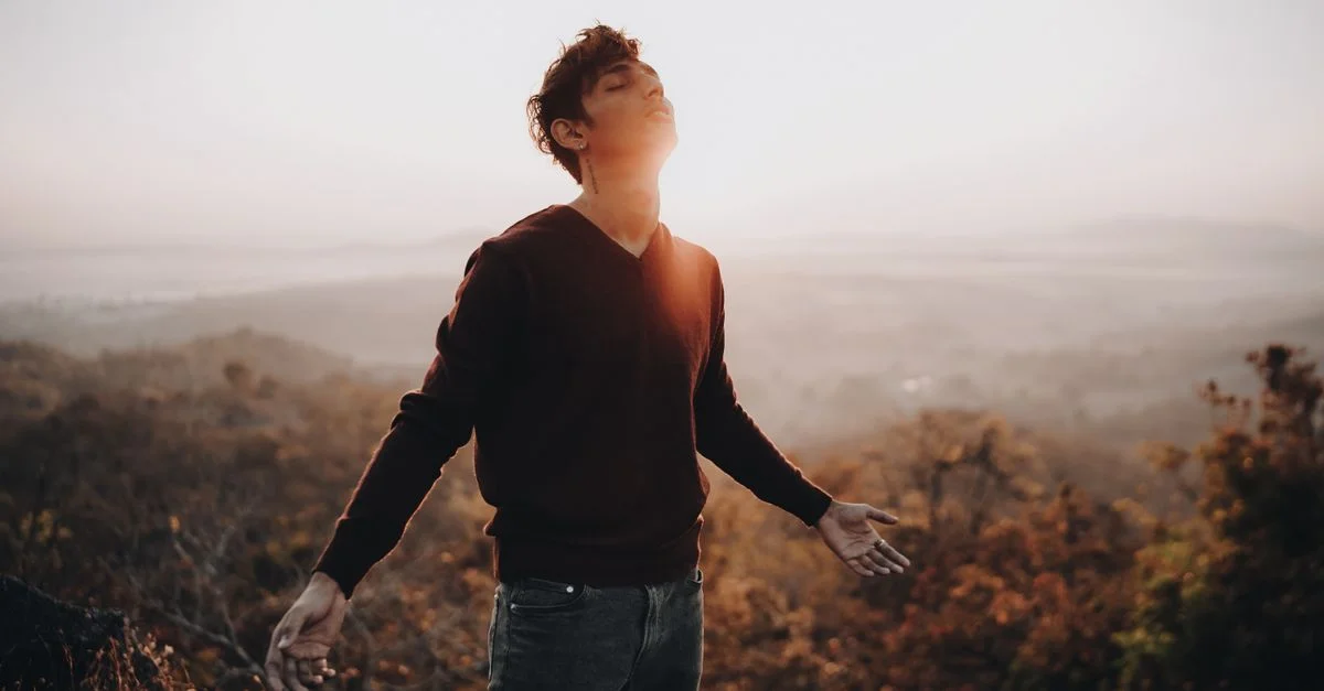 Un jeune homme debout sur une colline brumeuse au lever du soleil, embrassant la lumière et la nature.