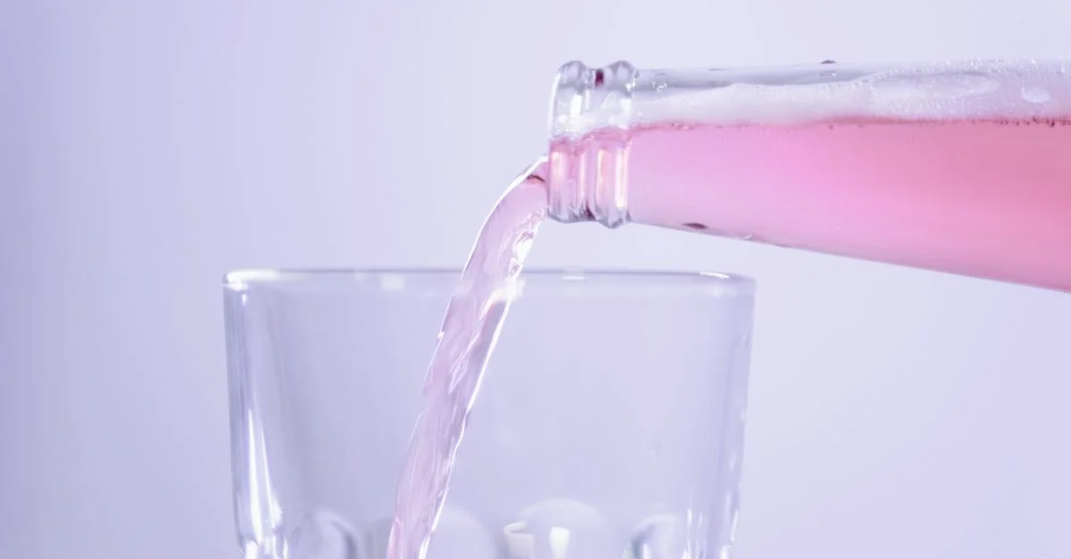 Gros plan sur une boisson rose versée d'une bouteille dans un verre clair sur fond blanc.