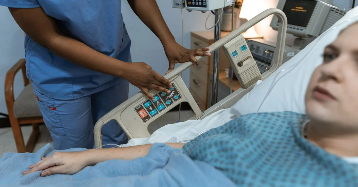 Une infirmière en tenue médicale aide un patient dans un lit d'hôpital avec des équipements médicaux à proximité.