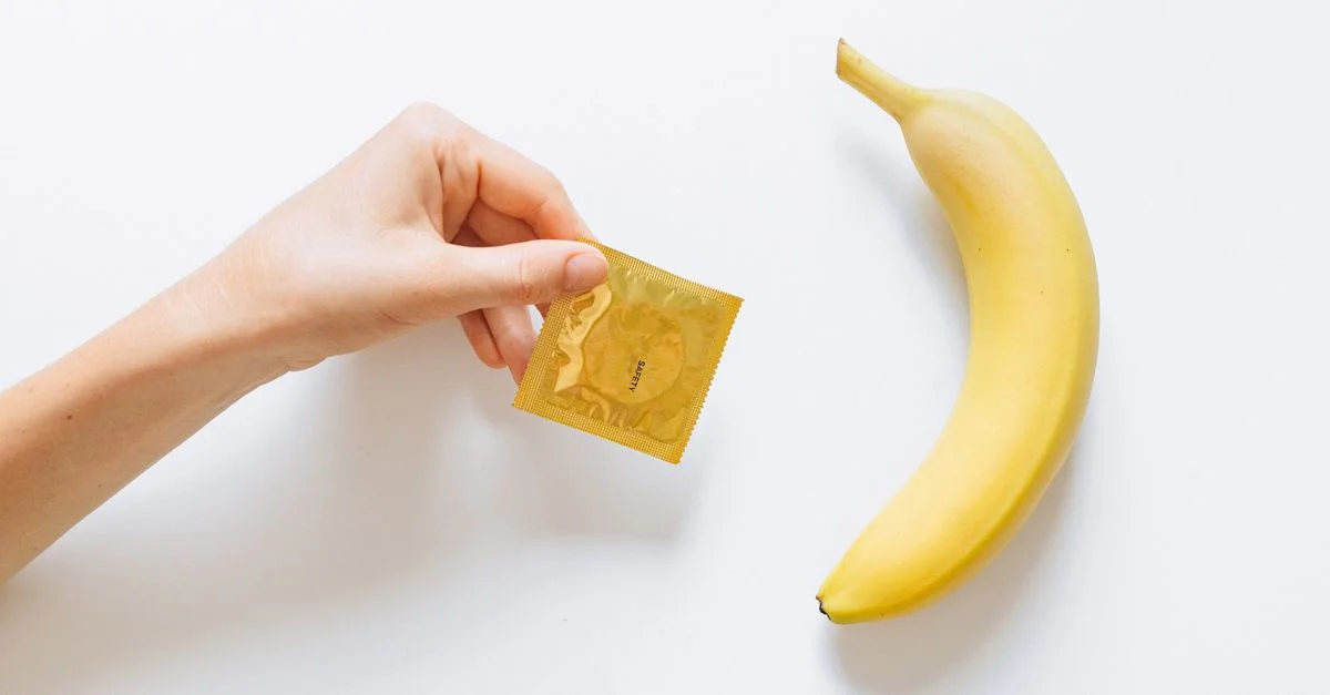 Une main tenant un préservatif à côté d'une banane sur un fond blanc, symbolisant l'éducation sur le sexe sécurisé.