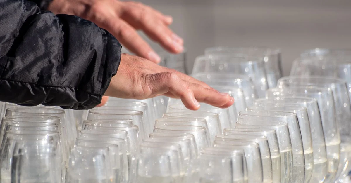Des mains jouent d'une harpe de verre faite de verres remplis d'eau, en plein soleil.