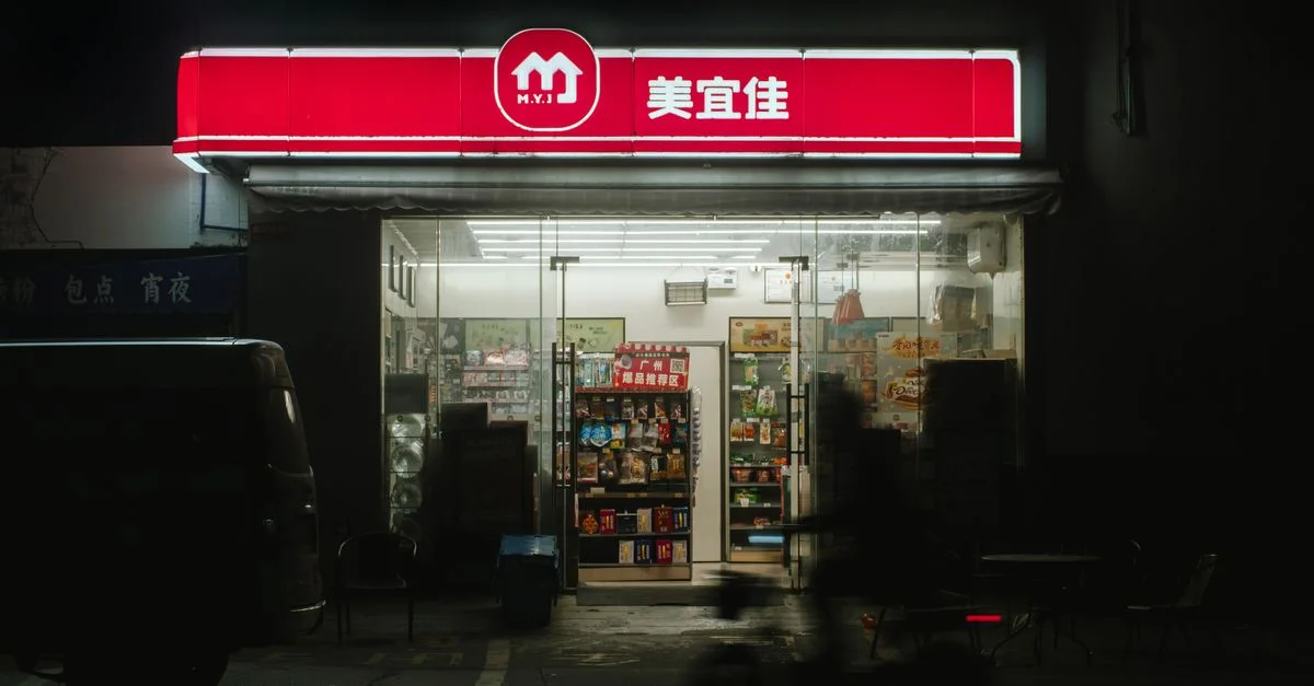 Un dépanneur faiblement éclairé avec une enseigne au néon lumineuse la nuit, doté de sièges extérieurs.
