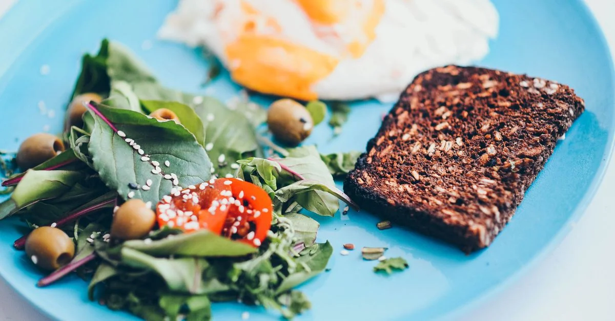 Une assiette de petit-déjeuner nutritif mettant en avant des œufs, du pain de seigle, une salade et des olives sur une assiette bleue vivante.
