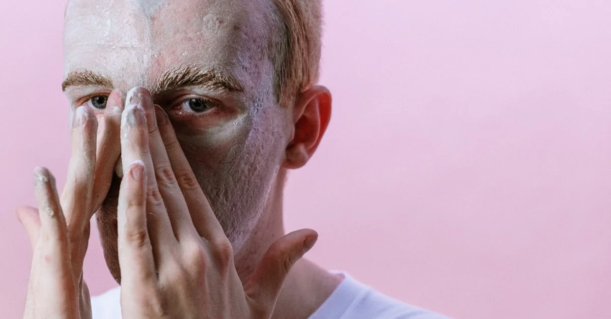 Jeune homme appliquant un masque en argile dans le cadre de sa routine de soins de la peau sur fond rose.