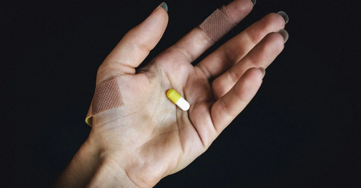 Prise de vue détaillée d'une main avec des pansements tenant une capsule jaune et blanche sur un fond sombre.