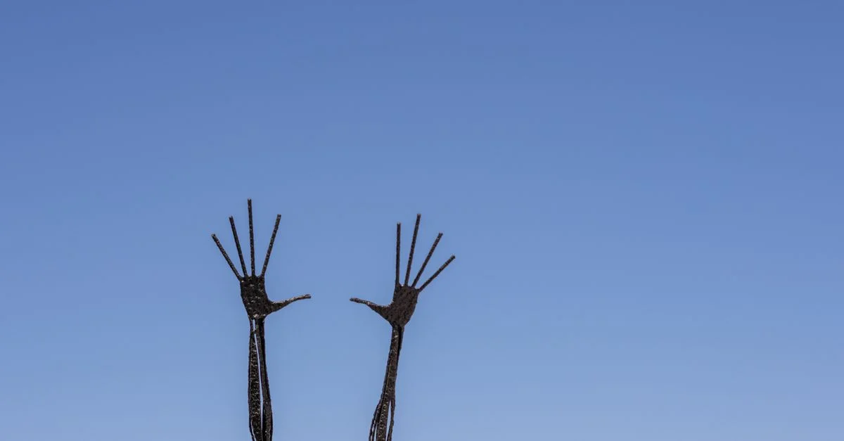 Sculpture métallique abstraite de mains s'élevant vers le ciel bleu clair, mettant en avant l'expression artistique.