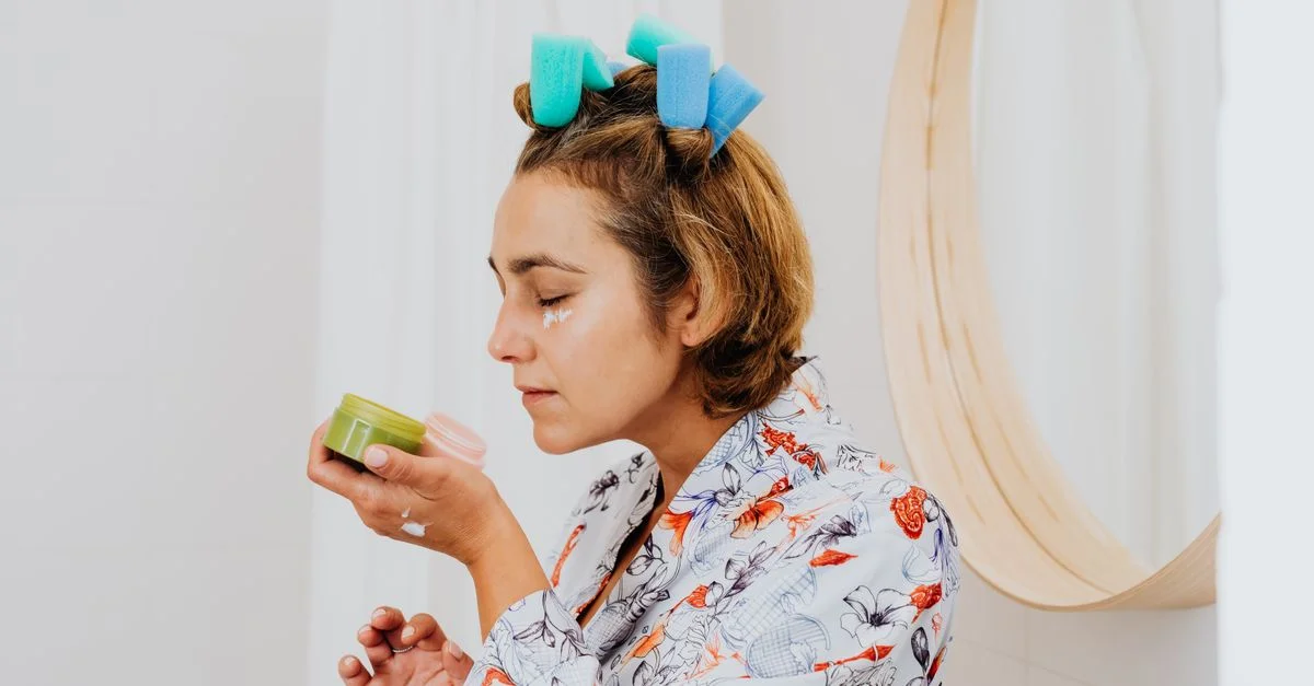 Femme avec des rouleaux dans les cheveux applique de la crème pour la peau devant un miroir de salle de bains.