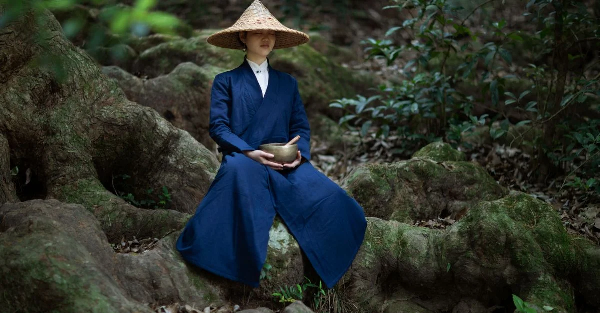 Une femme asiatique méditant dans une forêt, portant un chapeau traditionnel et tenant un bol.